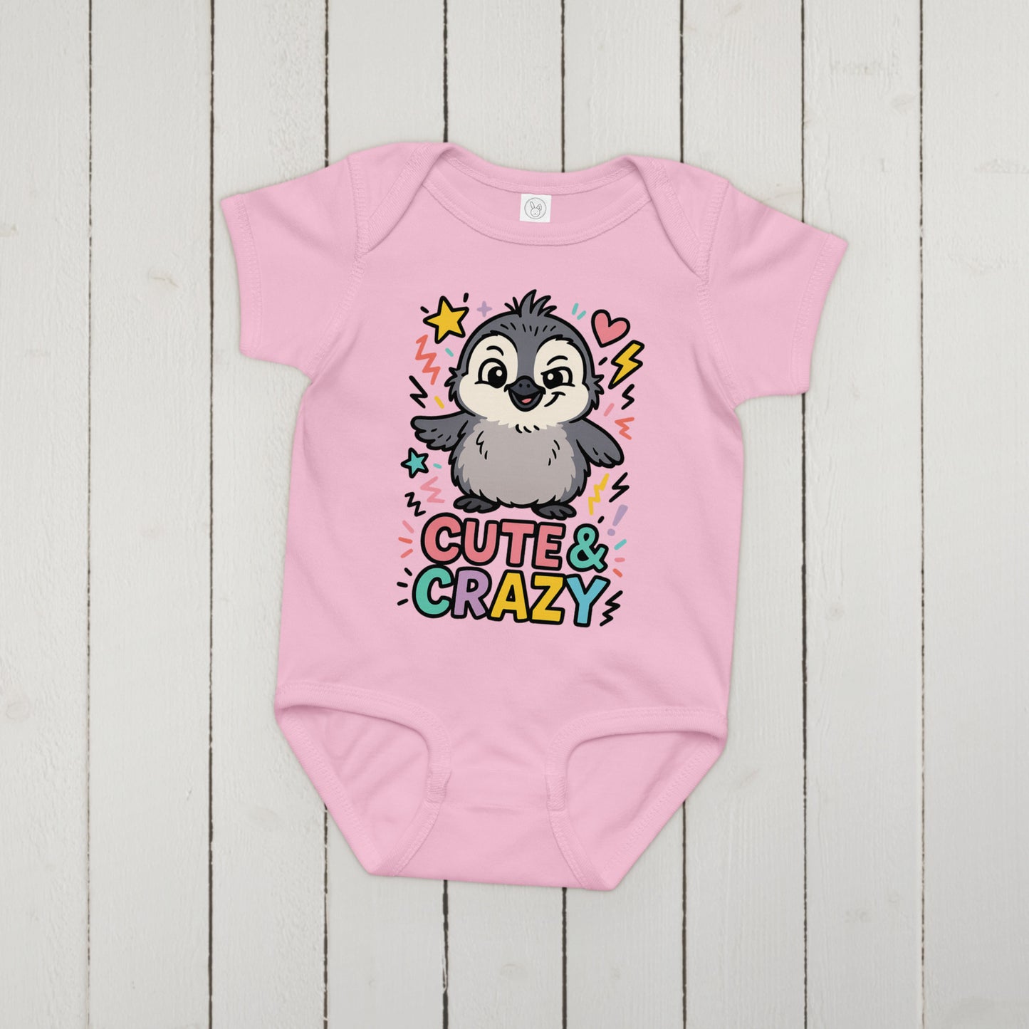 Pink baby onesie with a penguin graphic and 'Cute & Crazy' text on a wooden surface