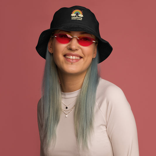 Person wearing a black bucket hat with a rainbow design and red sunglasses against a pink background