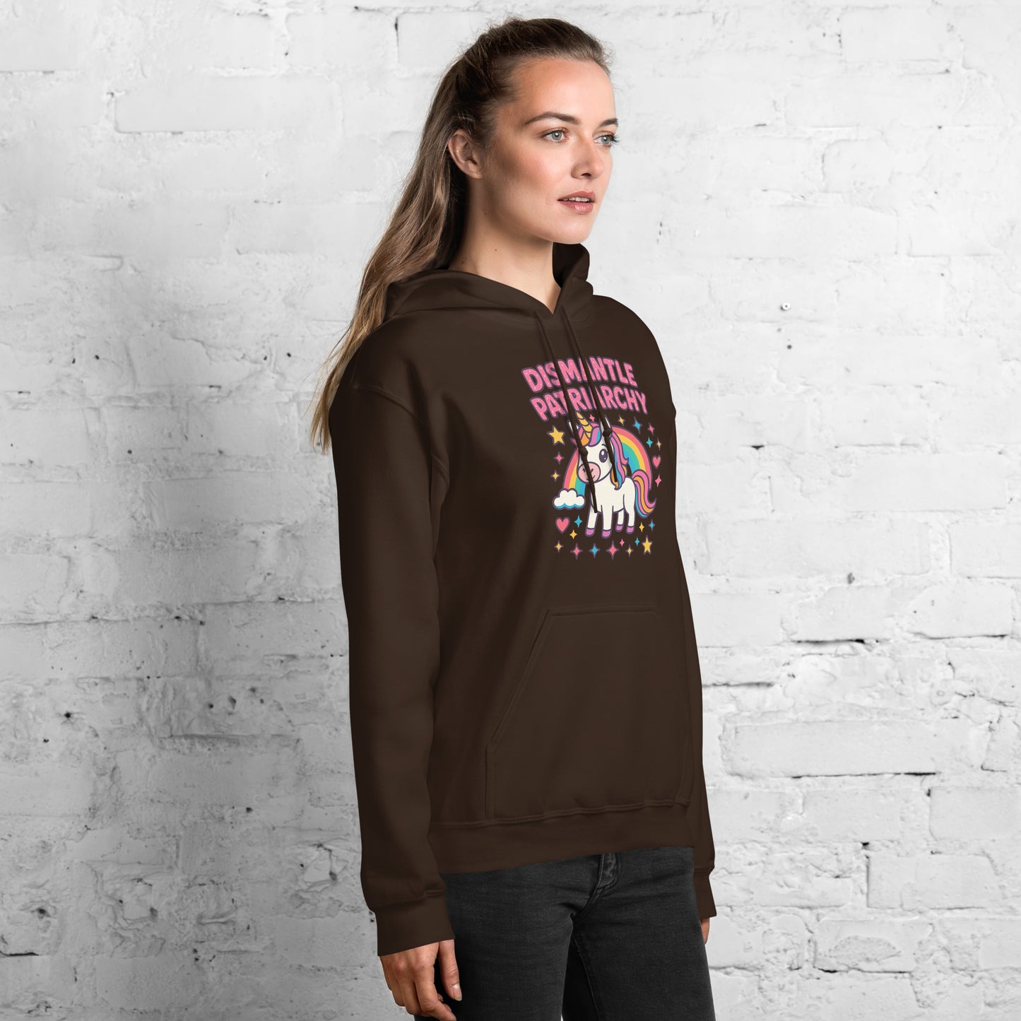 Woman wearing a brown hoodie with a colorful graphic and text against a white brick wall.