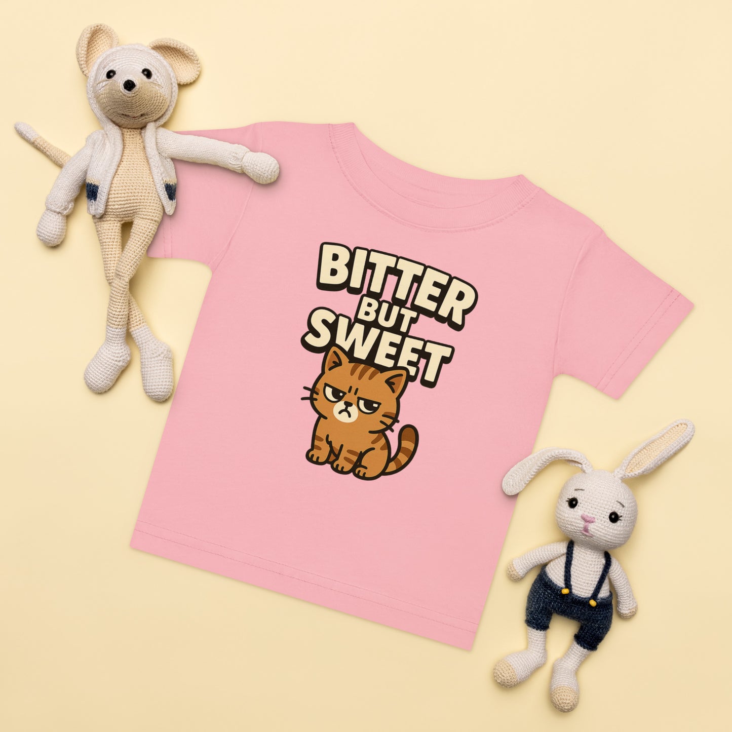 Pink t-shirt with a cat graphic and 'Bitter But Sweet' text, surrounded by two plush toys on a yellow background.