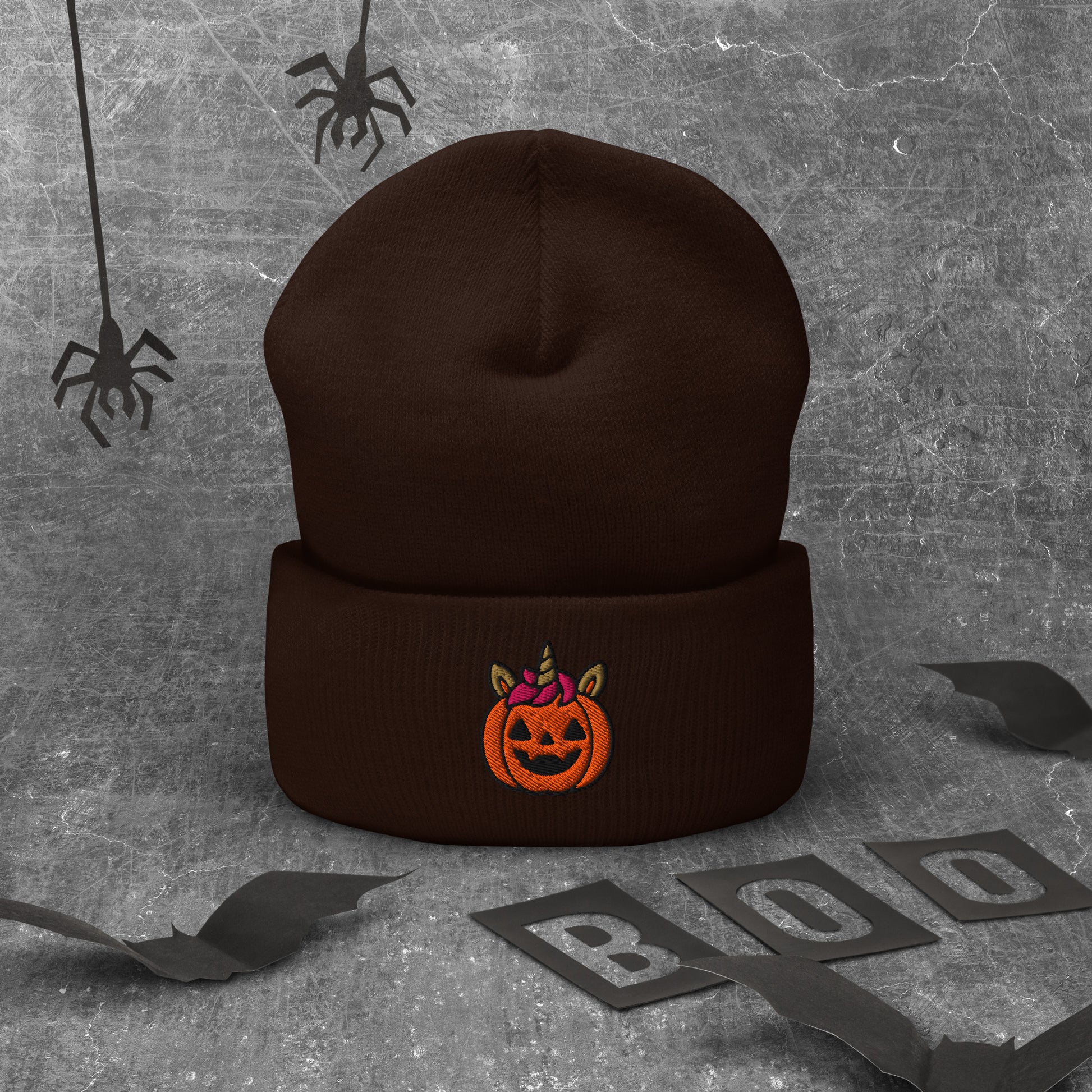 Brown beanie with Halloween pumpkin design on a textured gray background with spider webs and 'BOO' letters.