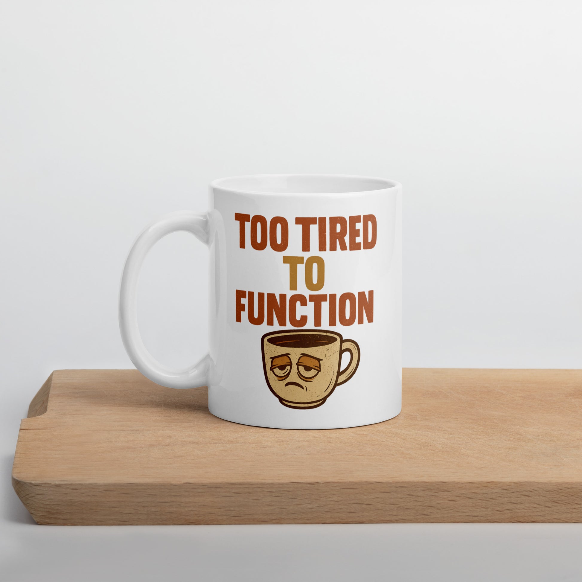 White mug with 'Too Tired to Function' text and coffee cup graphic on a wooden surface.