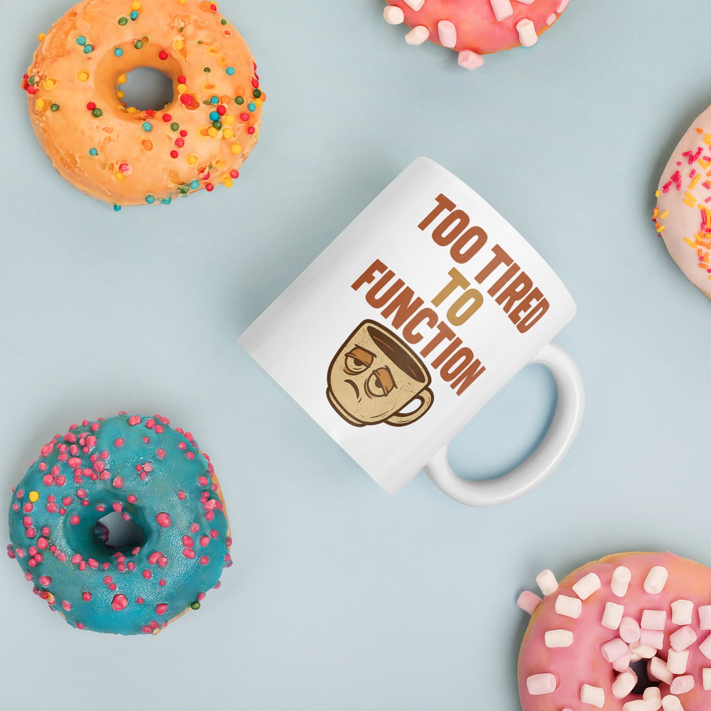 White mug with 'Too Tired to Function' text and coffee cup graphic, surrounded by colorful donuts on a light blue background