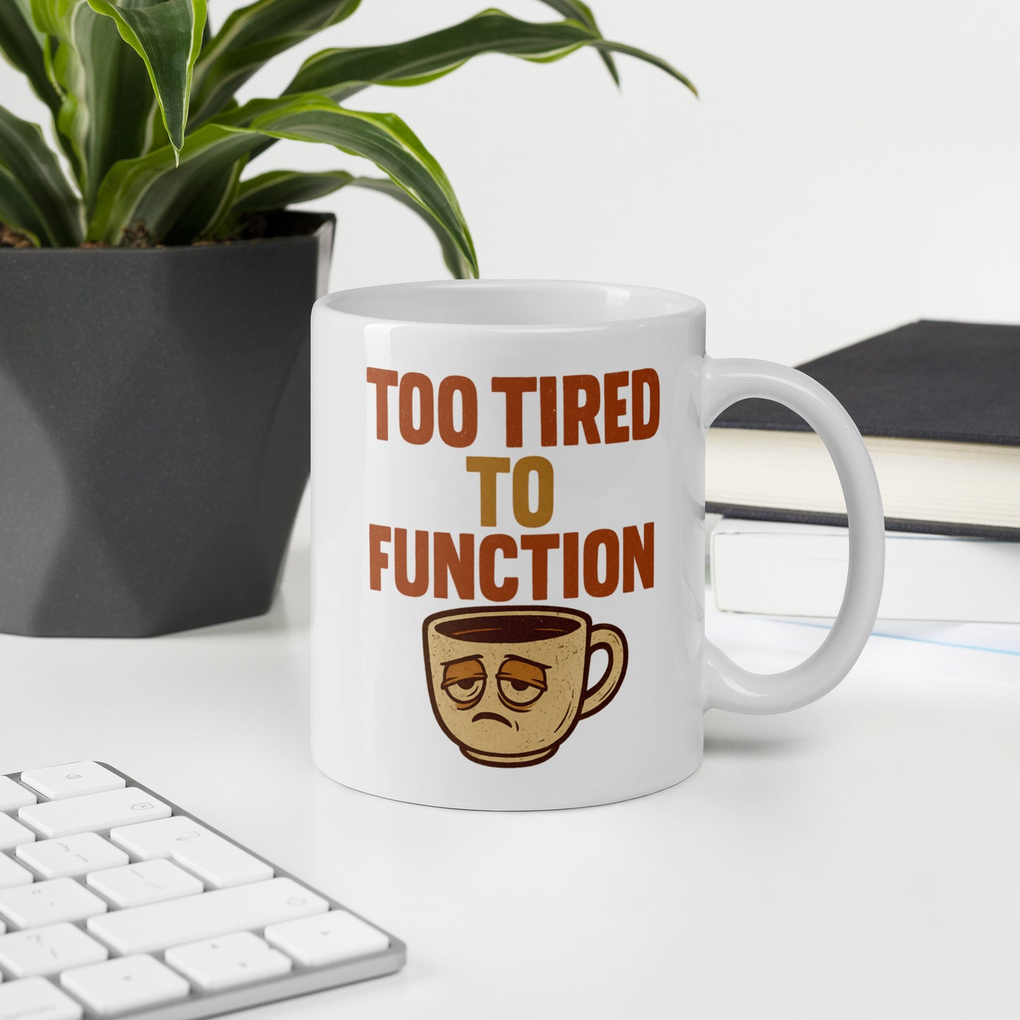 Mug with 'Too Tired to Function' text and coffee cup graphic on a desk with a plant and books in the background.