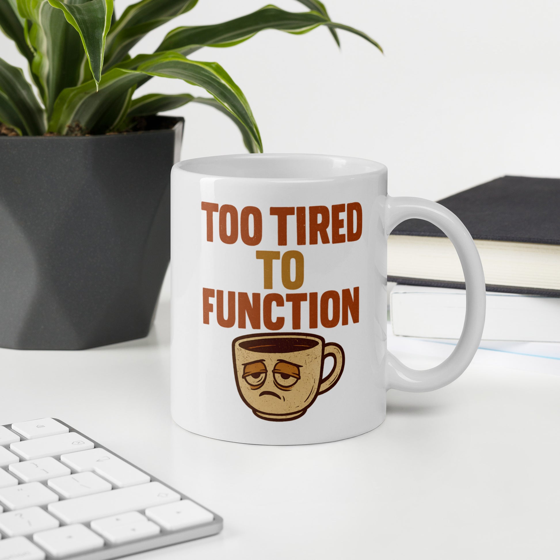 Mug with 'Too Tired to Function' text and coffee cup graphic on a desk with a plant and books in the background.