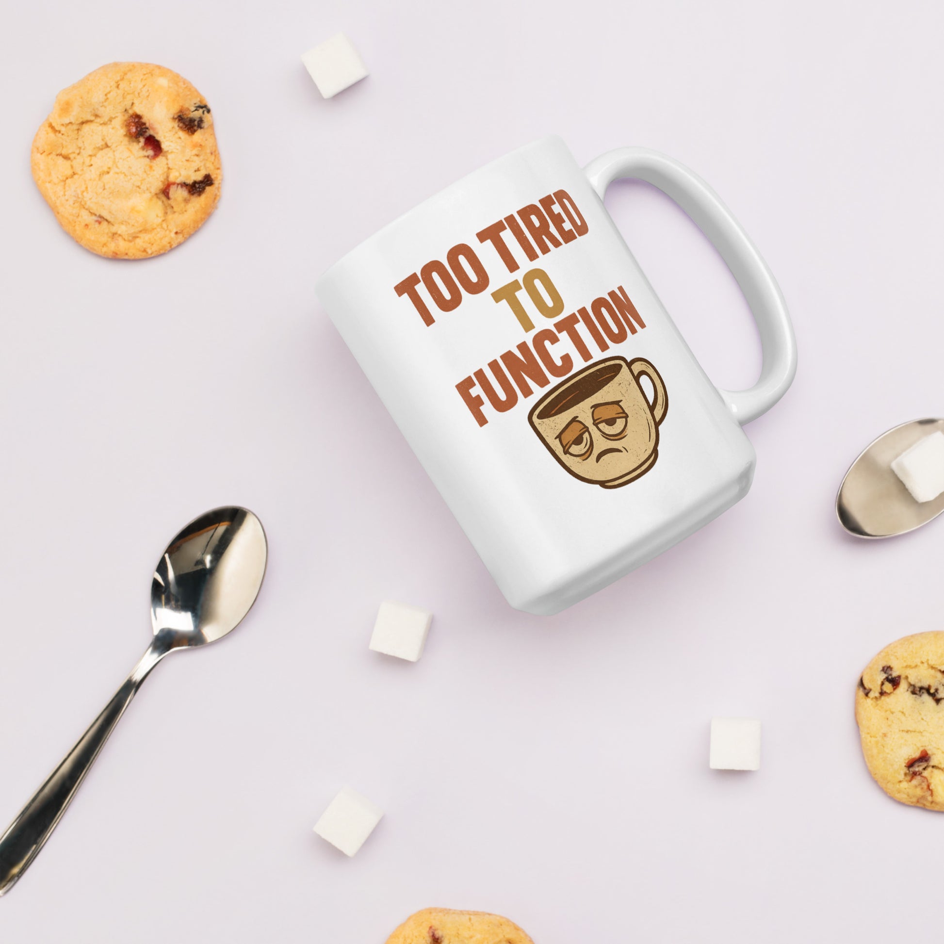 White mug with 'Too Tired to Function' text and coffee cup icon on a light background with cookies and a spoon.