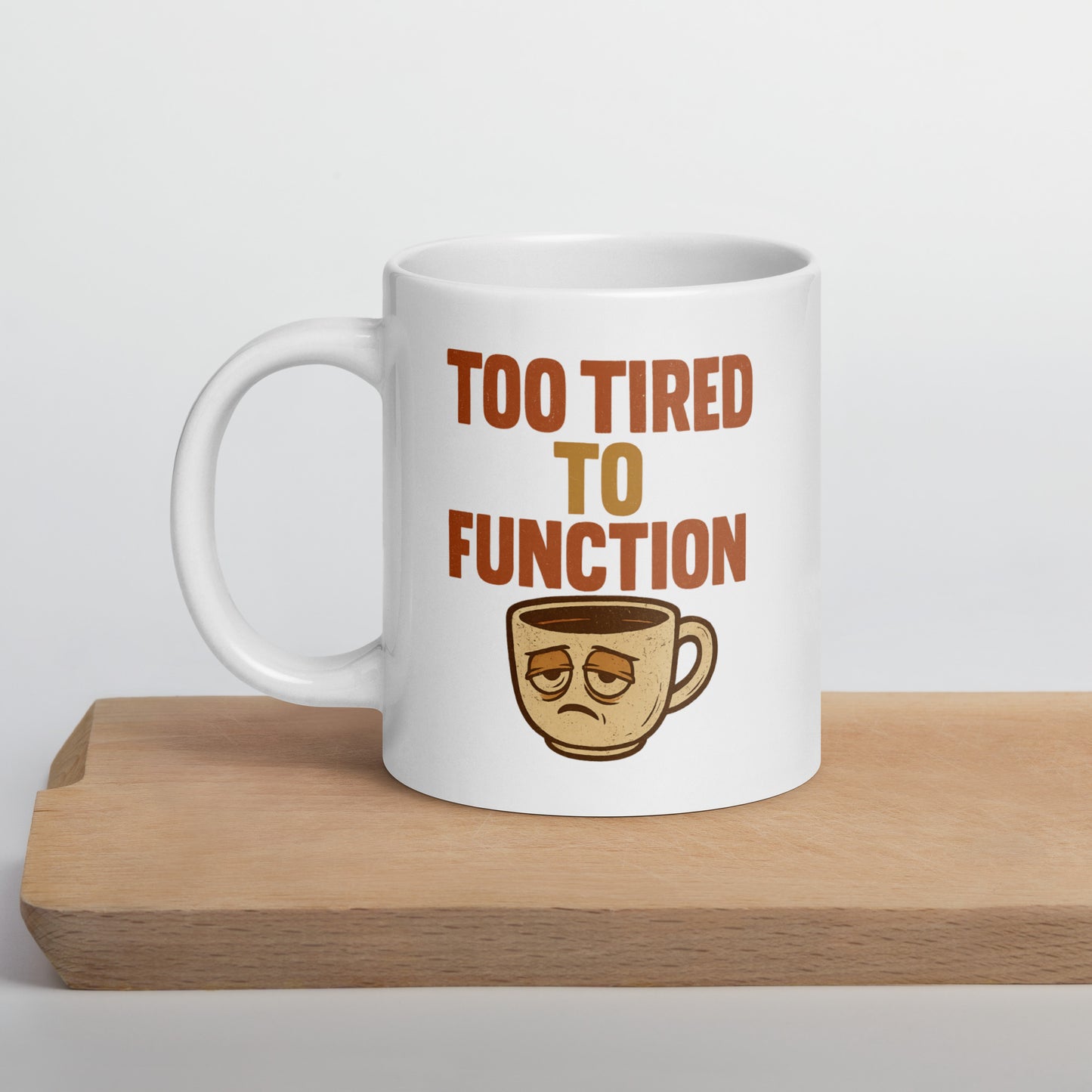 White mug with 'Too Tired to Function' text and coffee cup graphic on a wooden surface.