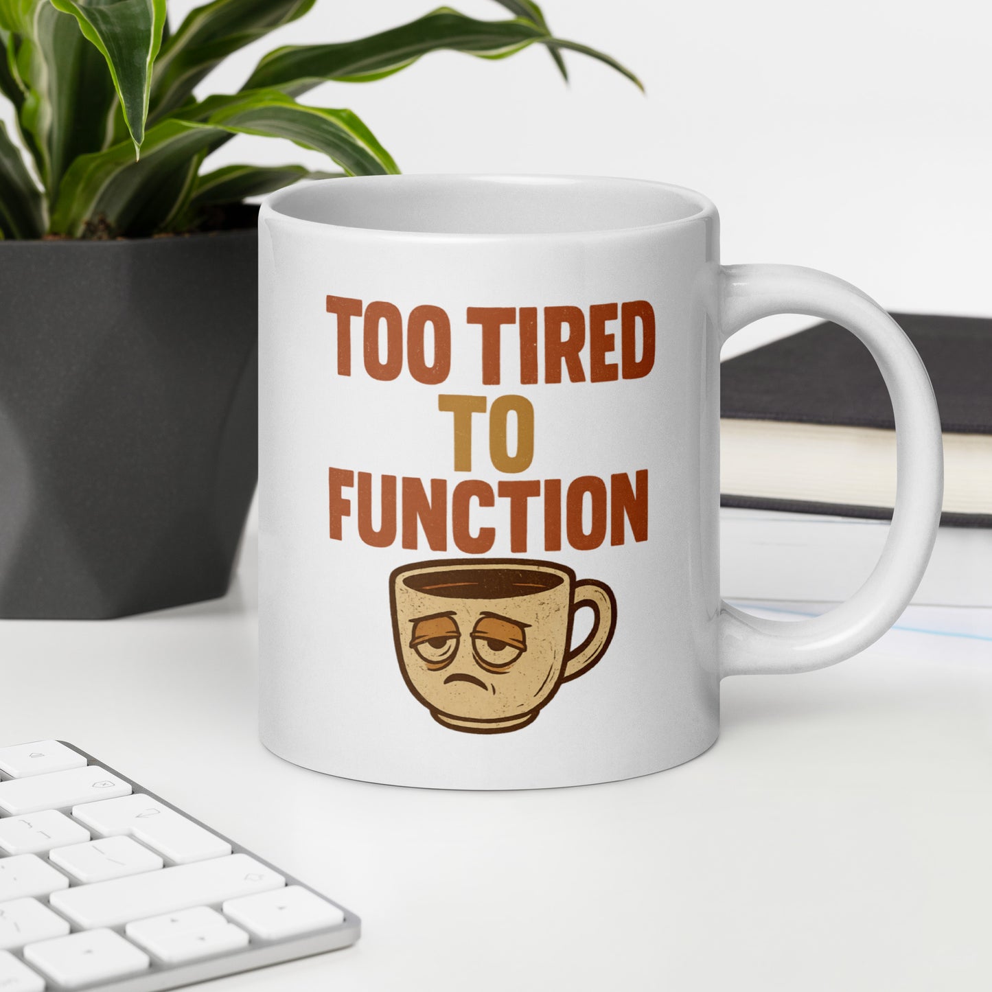 White mug with 'Too Tired to Function' text and coffee cup graphic on a desk with a plant and books in the background.