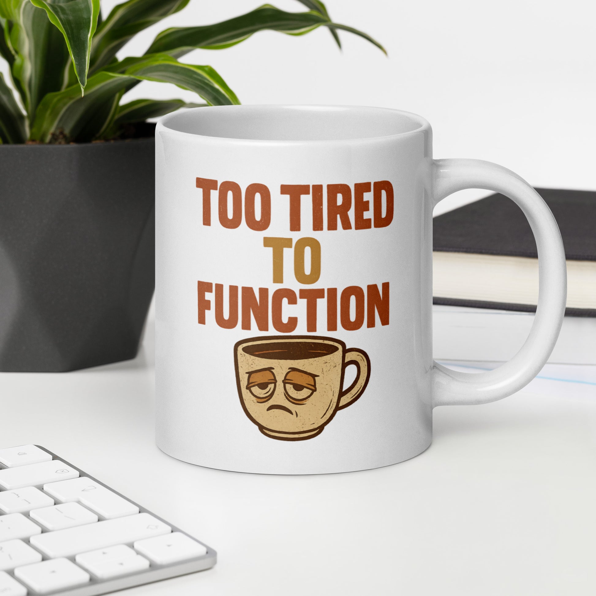 White mug with 'Too Tired to Function' text and coffee cup graphic on a desk with a plant and books in the background.